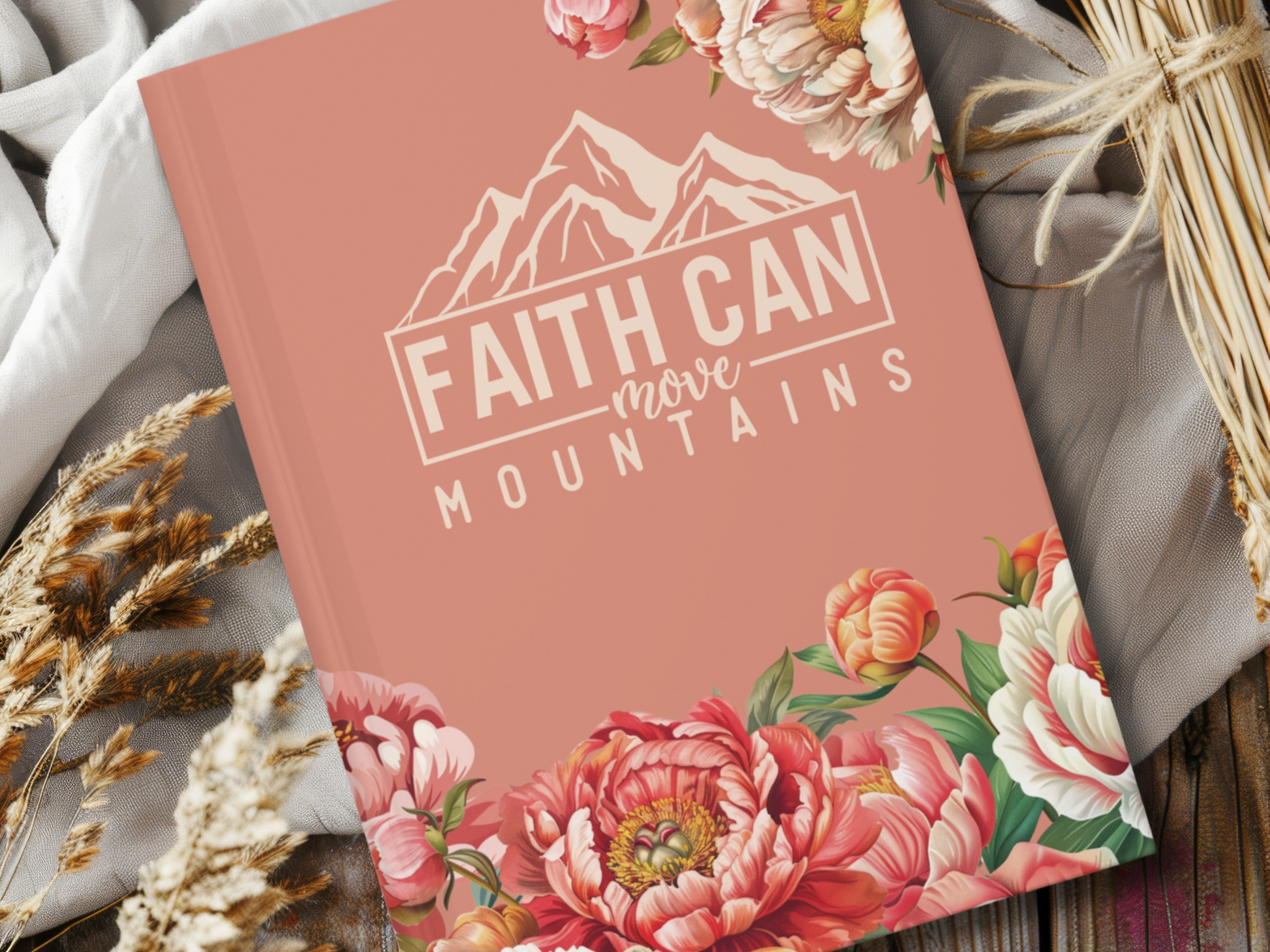 Pink journal cover with floral decor, featuring the text "Faith Can Move Mountains" and a mountain outline. The background has wheat stalks and light fabric, conveying a hopeful, serene tone.
