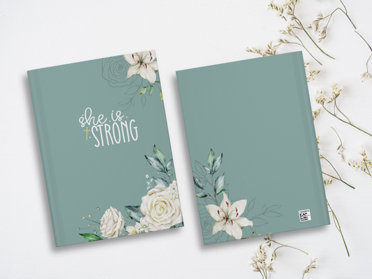 Two teal notebooks lie on a white surface, adorned with elegant white floral designs. One cover reads "She is Strong" in white script. Delicate flowers surround.