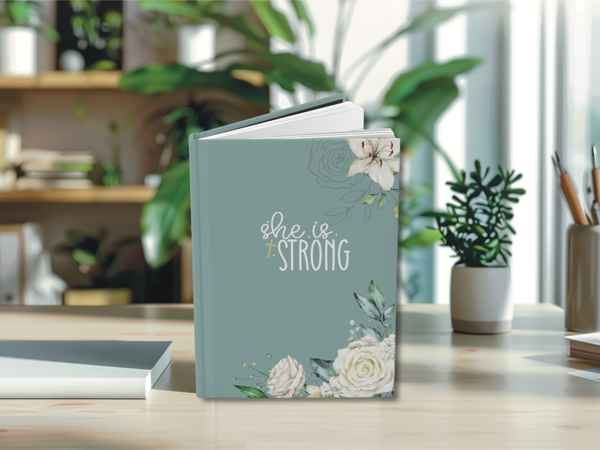A teal journal on a desk titled "She is Strong," adorned with white flowers. The background features blurred shelves and greenery, creating a serene atmosphere.