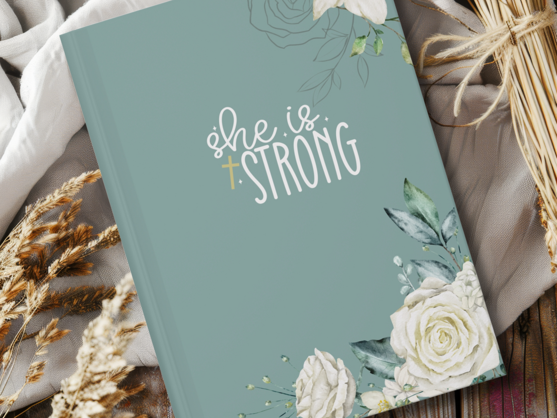 A teal religious journal with "She is Strong" in white script, adorned with floral designs. It's set on fabric with dried wheat, conveying empowerment and nature.