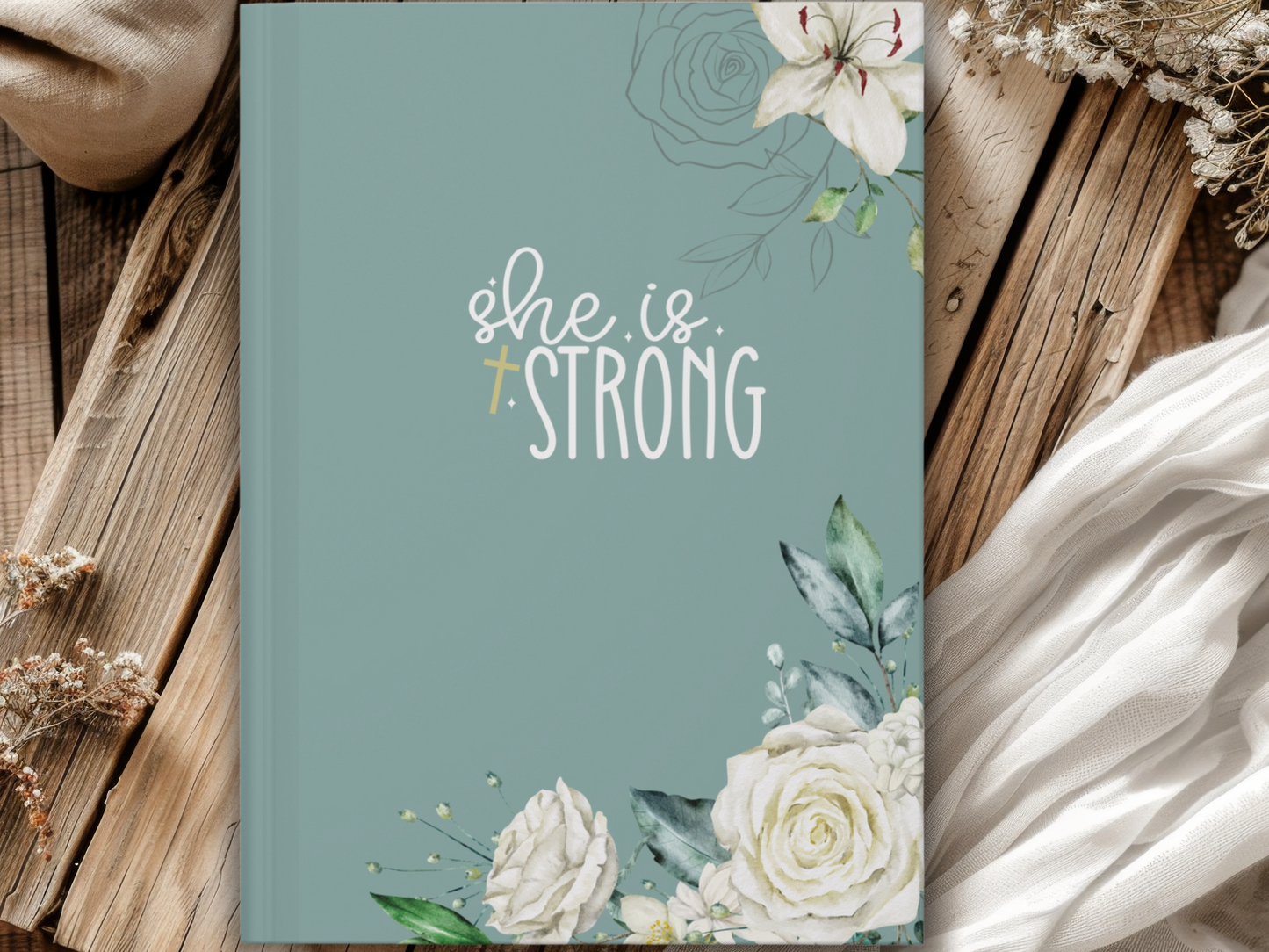 A pastel teal notebook with "She is Strong" in white, adorned with floral designs of white roses and leaves. It rests on rustic wooden planks and a white fabric, creating a calm, uplifting atmosphere.