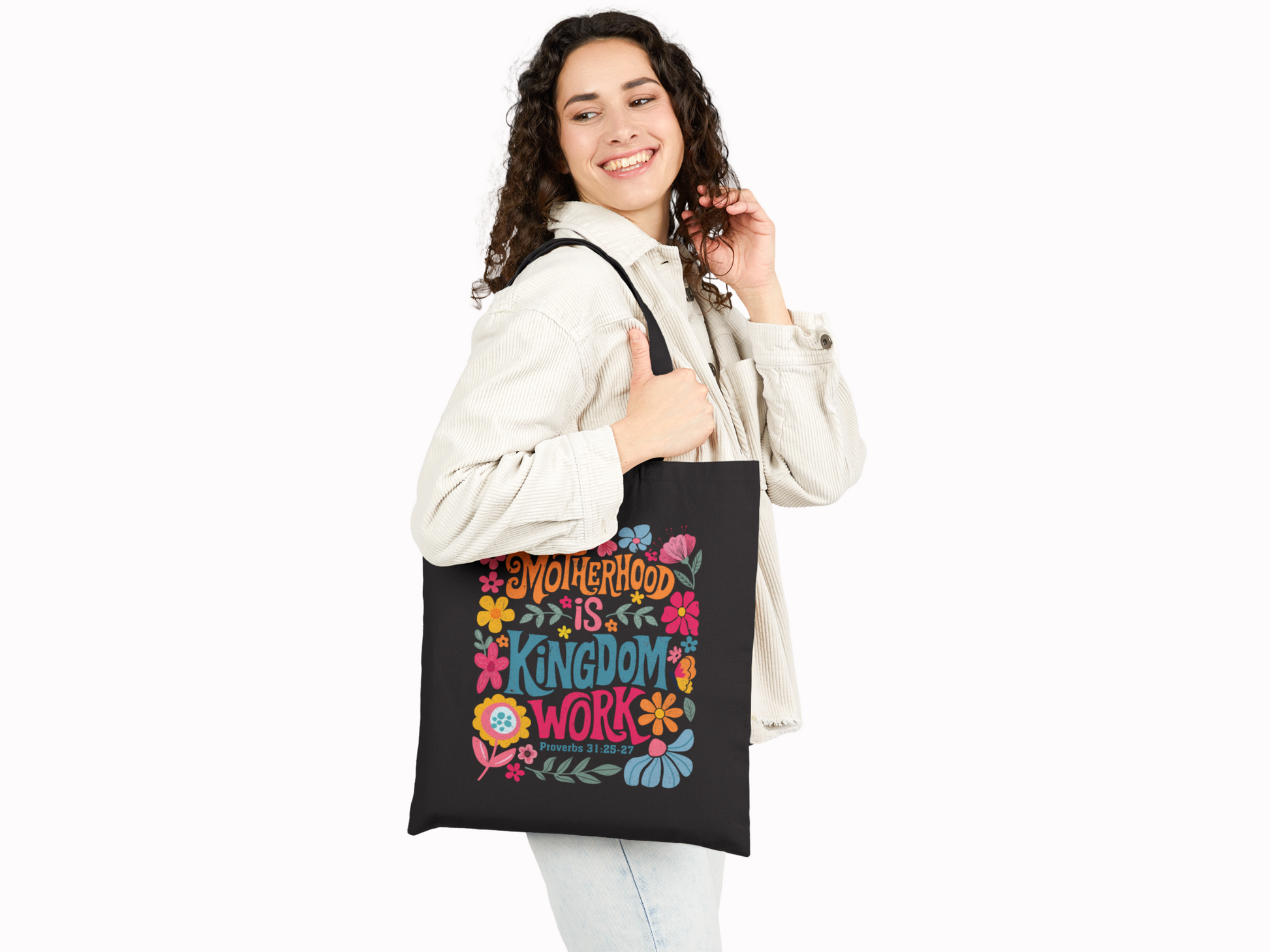 A smiling woman with curly hair holds a black tote bag featuring colorful flowers and the text "Motherhood is Kingdom Work" in vibrant letters.