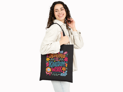A smiling woman with curly hair holds a black tote bag featuring colorful flowers and the text "Motherhood is Kingdom Work" in vibrant letters.