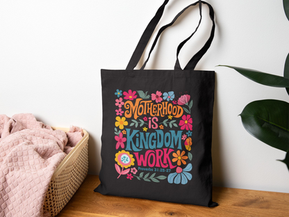 A black tote bag with colorful floral graphics and the words "Motherhood is Kingdom Work" rests on a wooden surface. A cozy pink blanket is in a basket nearby.