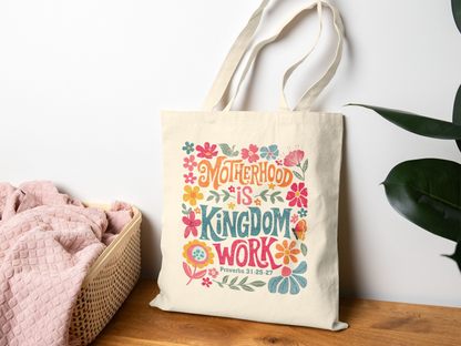 A beige tote bag with colorful floral designs and the phrase "Motherhood is Kingdom Work" hangs on a wall. A basket with a pink blanket is nearby.