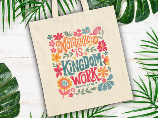 Canvas tote bag with colorful floral design and text "Motherhood is Kingdom Work, Proverbs 31:25-27." Surrounded by green palm leaves on light wood.