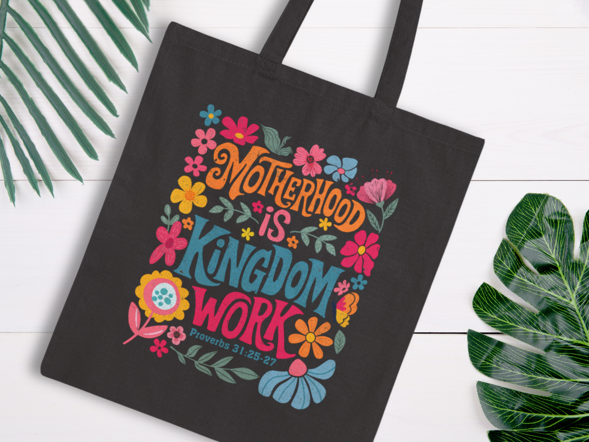 A black tote bag with colorful floral designs and bold text reading "Motherhood is Kingdom Work, Proverbs 31:25-27," against a light background. Bright, cheerful tone.