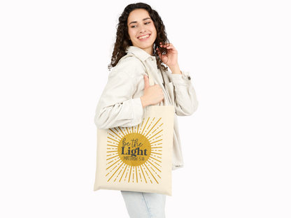 Smiling woman with curly hair holding a beige tote bag. Bag features a sunburst design with the text "Be the Light," creating an uplifting mood.