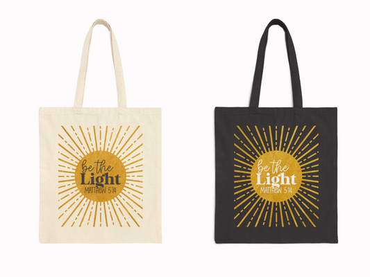 Two tote bags, one beige and one black, feature a yellow sunburst design with the words "be the Light Matthew 5:14" in the center, conveying inspiration.