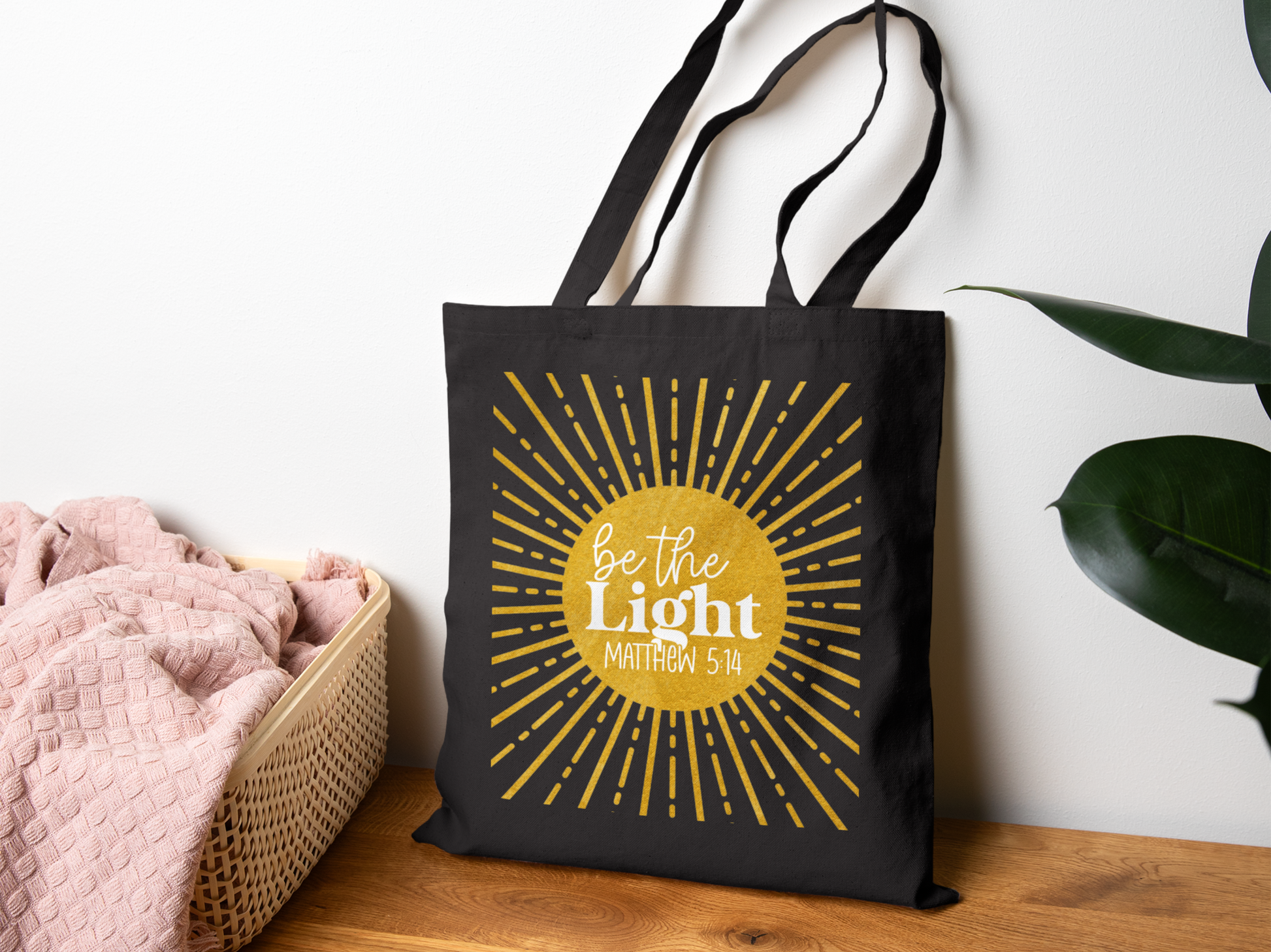 A black tote bag with "Be the Light, Matthew 5:14" in white text over a yellow sunburst design. It hangs on a wall, next to a basket with a pink blanket.