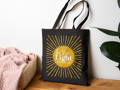 A black tote bag with "Be the Light, Matthew 5:14" in white text over a yellow sunburst design. It hangs on a wall, next to a basket with a pink blanket.