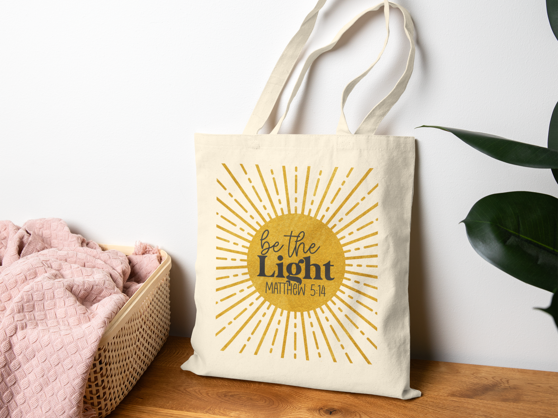 Canvas tote bag with "Be the Light, Matthew 5:14" and sunburst design, sits on a wooden surface near a basket with pink fabric and a green plant.