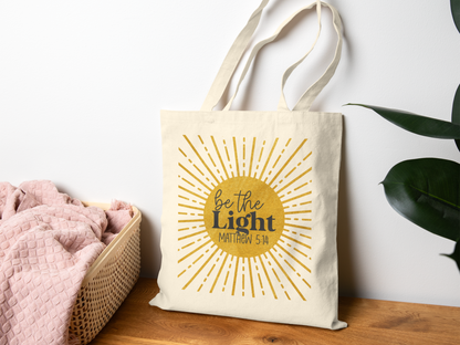 Canvas tote bag with "Be the Light, Matthew 5:14" and sunburst design, sits on a wooden surface near a basket with pink fabric and a green plant.