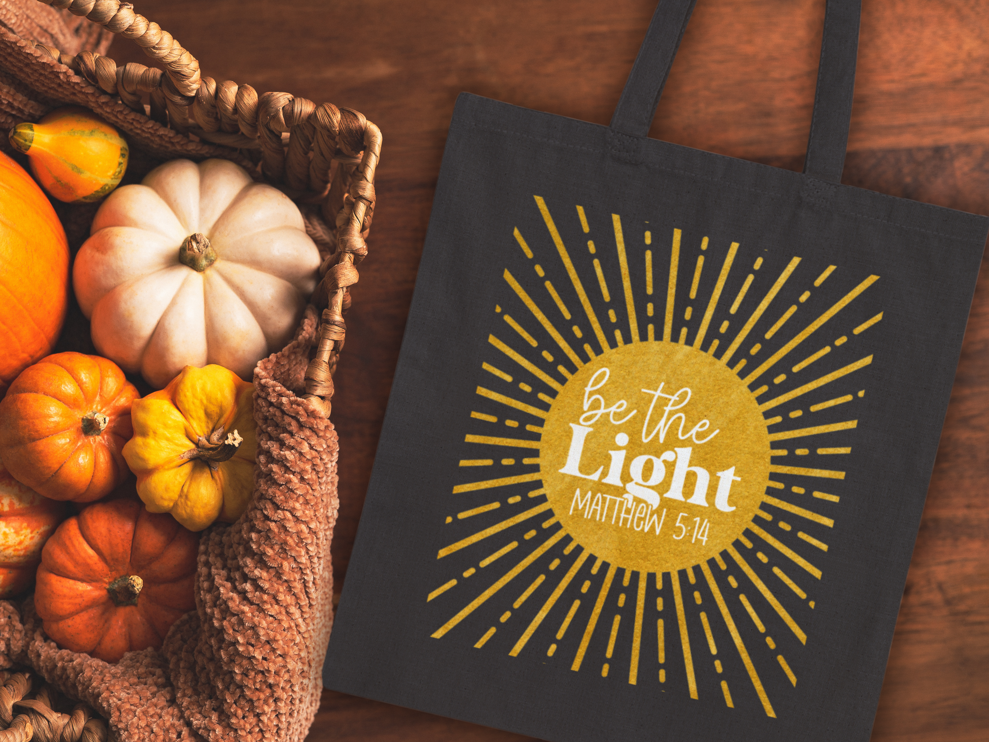 Black bag with "Be the Light, Matthew 5:14" text inside a sunburst design. Next to it, a basket holds small pumpkins on a wooden surface. Autumn mood.