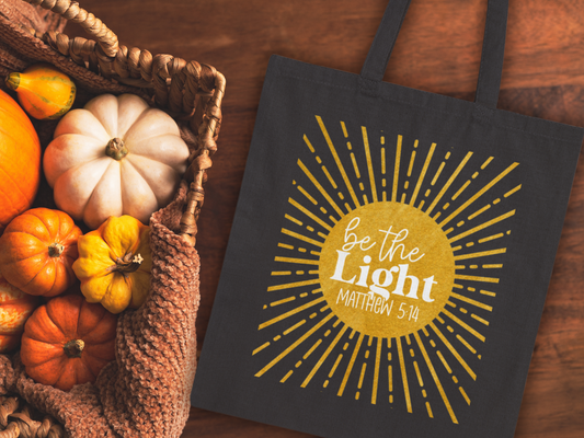 Black bag with "Be the Light, Matthew 5:14" text inside a sunburst design. Next to it, a basket holds small pumpkins on a wooden surface. Autumn mood.