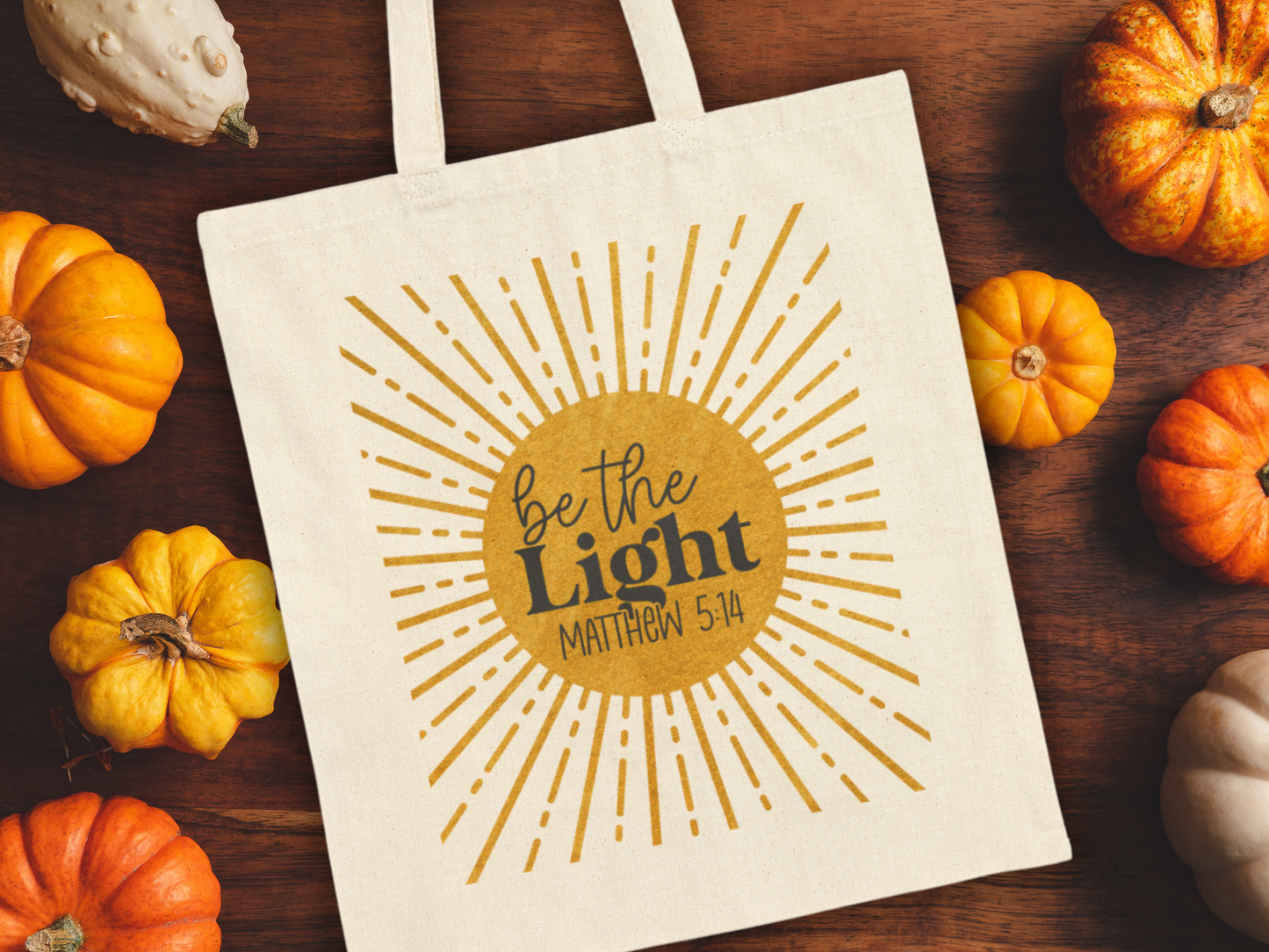 A canvas bag on a wooden table, surrounded by small pumpkins. The bag features a sunburst design with "Be the Light, Matthew 5:14" in bold letters.
