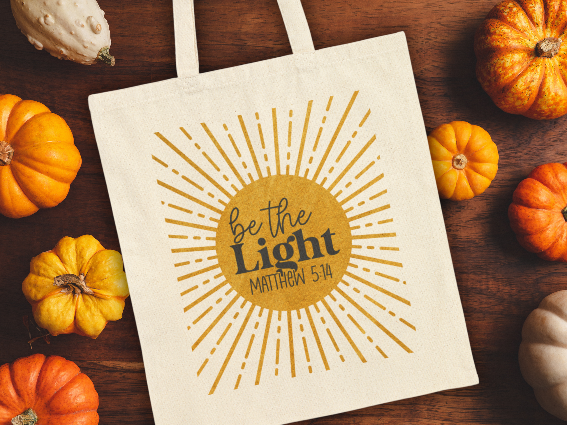 A canvas bag on a wooden table, surrounded by small pumpkins. The bag features a sunburst design with "Be the Light, Matthew 5:14" in bold letters.