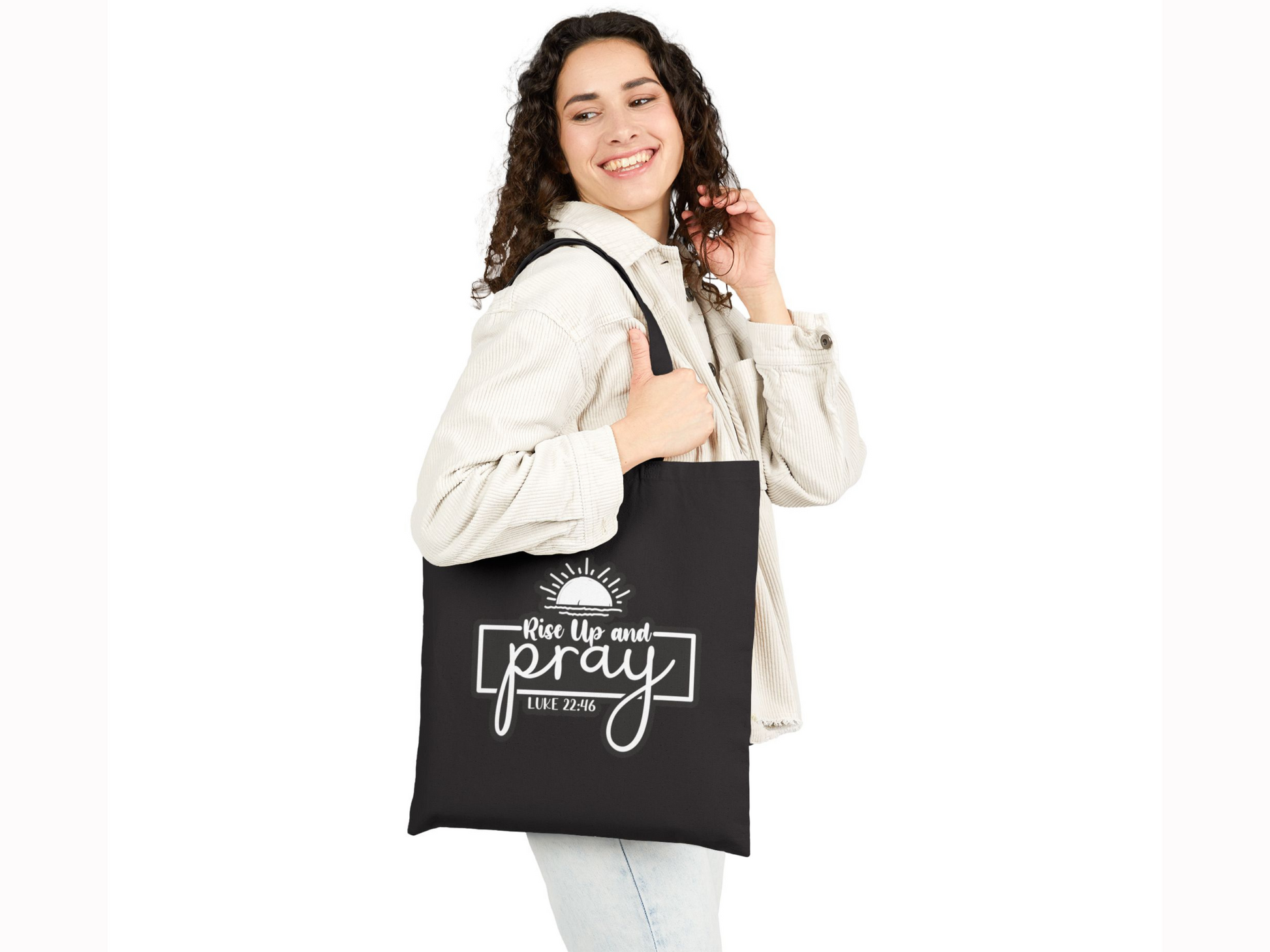 Smiling woman with curly hair holds a black tote bag that reads "Rise Up and Pray, Luke 22:46" featuring a sun graphic. She wears a light shirt and jeans.