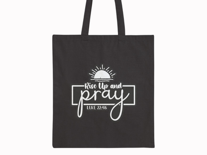 Black tote bag featuring the text "Rise Up and Pray" with a sunrise graphic above, and "Luke 22:46" below in white. Simple and inspirational design.