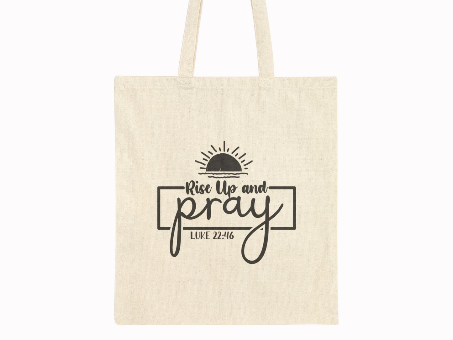 Beige tote bag with black text "Rise Up and Pray" beneath a half-sun graphic. The phrase "Luke 22:46" is displayed below, conveying a spiritual tone.