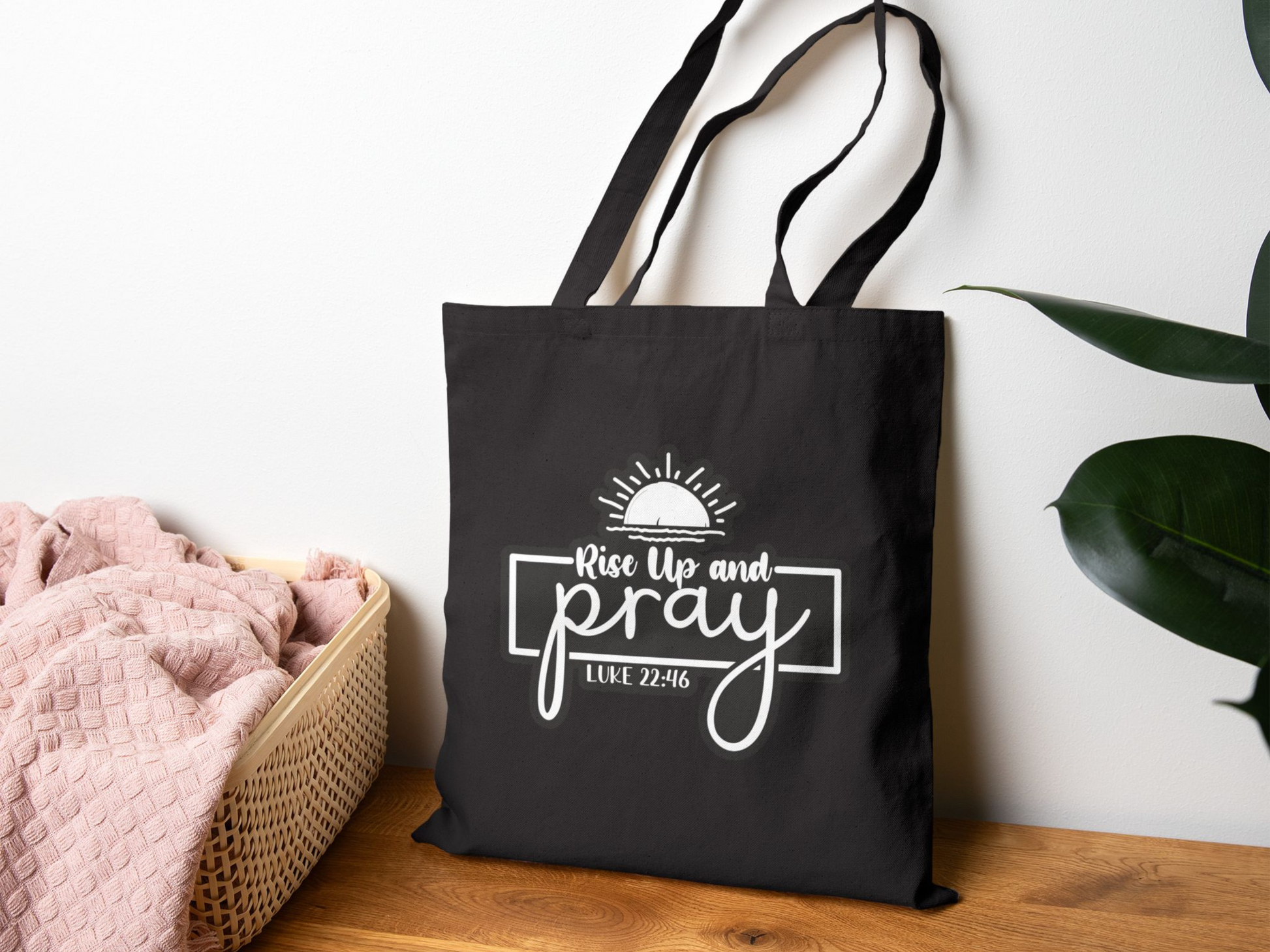 Black tote bag with the words "Rise Up and Pray Luke 22:46" in white, featuring a sun design. It's hanging by a basket of pink towels on a wooden surface. Calm atmosphere.