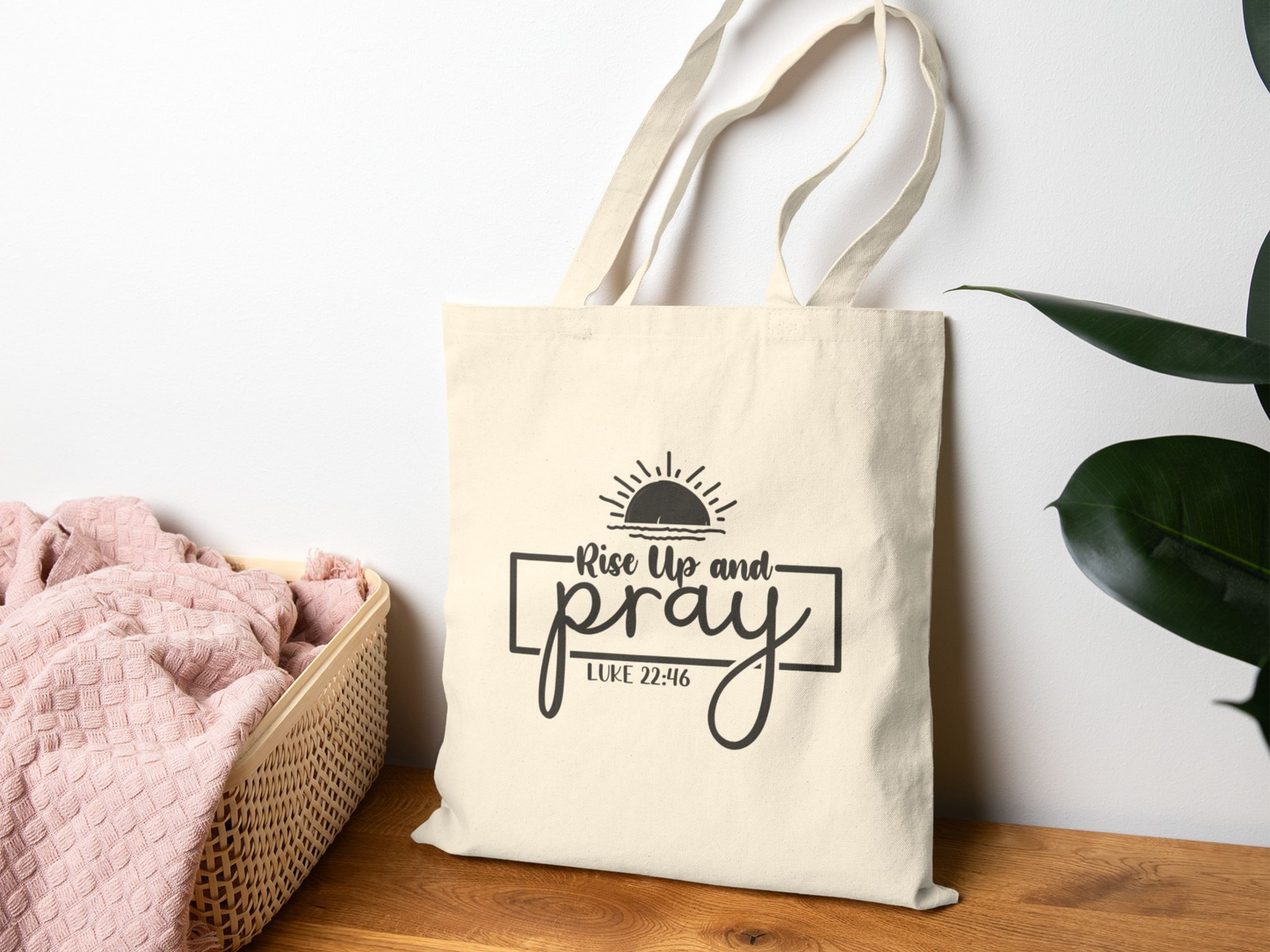 A beige tote bag with "Rise Up and Pray" and a sun graphic sits on a wooden surface. Nearby is a pink blanket in a basket and a large green plant.
