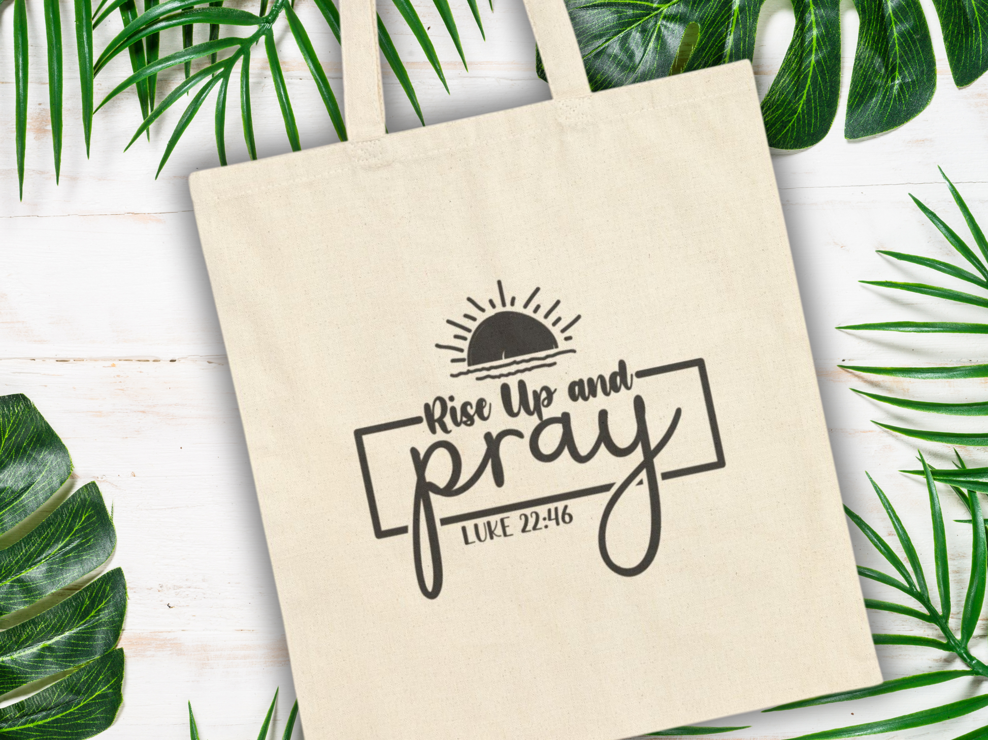 A beige tote bag with the words "Rise Up and Pray, Luke 22:46" in black, beneath a rising sun graphic. Surrounded by green tropical leaves on a white wooden surface.