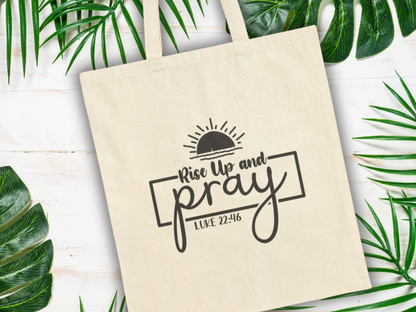 A beige tote bag with the words "Rise Up and Pray, Luke 22:46" in black, beneath a rising sun graphic. Surrounded by green tropical leaves on a white wooden surface.