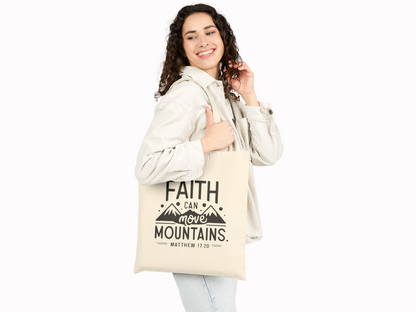 A smiling person with curly hair holds a beige bag featuring the text "Faith can move mountains." They wear a light corduroy jacket, conveying positivity.