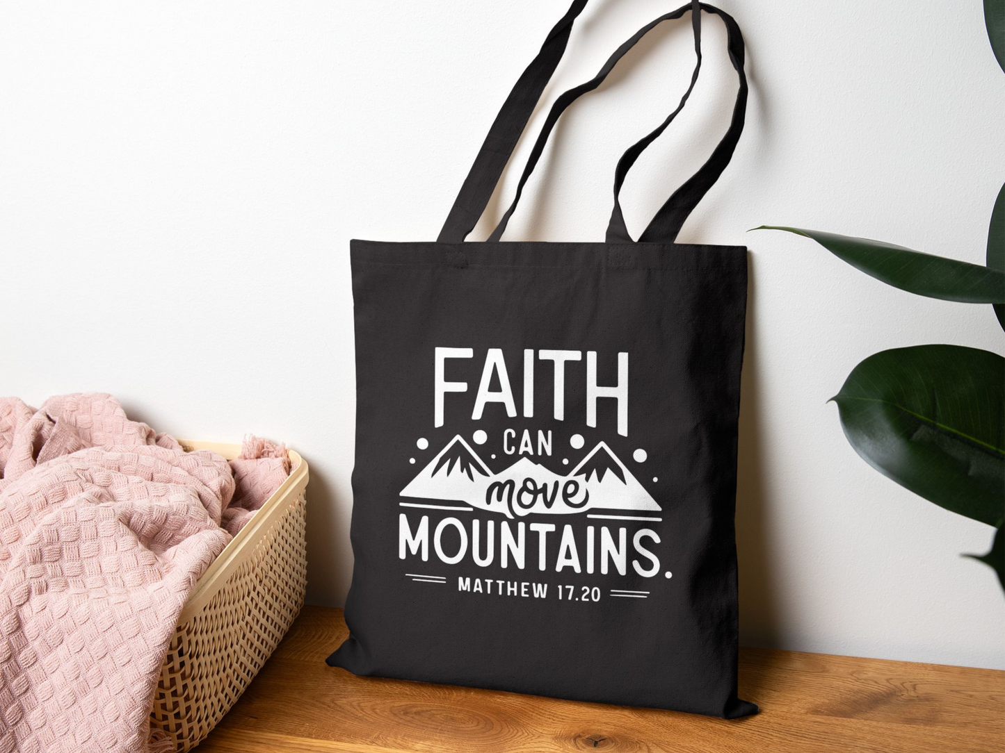 A black tote bag with the phrase "Faith Can Move Mountains, Matthew 17:20" in white text, featuring mountain graphics. It hangs on a wall near a pink blanket in a basket, with a green plant on the right, conveying a calm, inspirational tone.