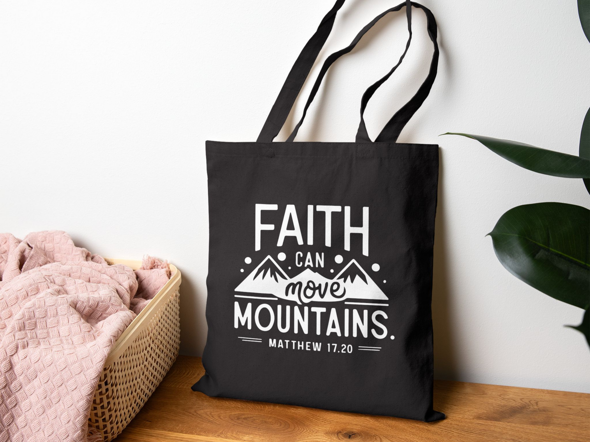 A black tote bag with the phrase "Faith Can Move Mountains, Matthew 17:20" in white text, featuring mountain graphics. It hangs on a wall near a pink blanket in a basket, with a green plant on the right, conveying a calm, inspirational tone.