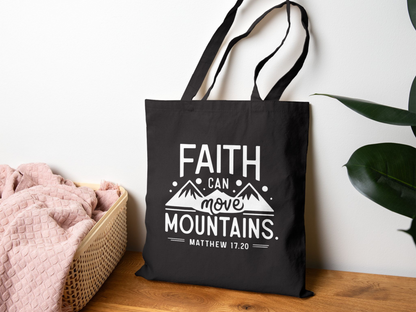 A black tote bag with the phrase "Faith Can Move Mountains, Matthew 17:20" in white text, featuring mountain graphics. It hangs on a wall near a pink blanket in a basket, with a green plant on the right, conveying a calm, inspirational tone.