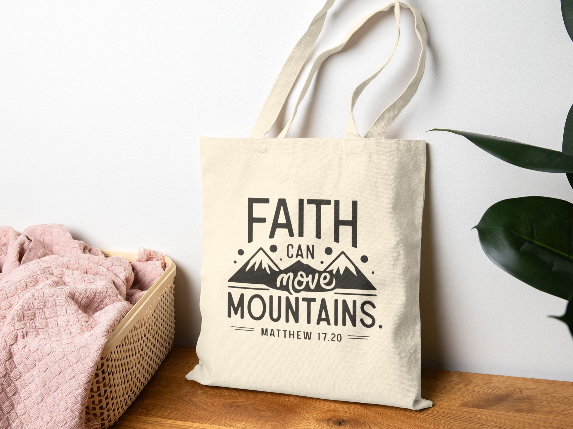 Beige bag with "Faith can move mountains. Matthew 17:20" text and mountain graphic, beside pink blanket in basket on wooden surface."