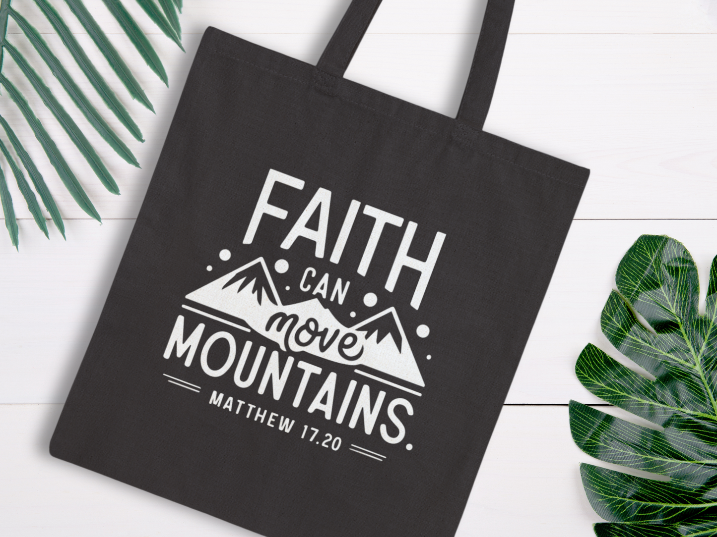 Black bag with white text "Faith can move mountains" and a mountain graphic. Surrounded by green leaves on a white wooden background. Uplifting tone.