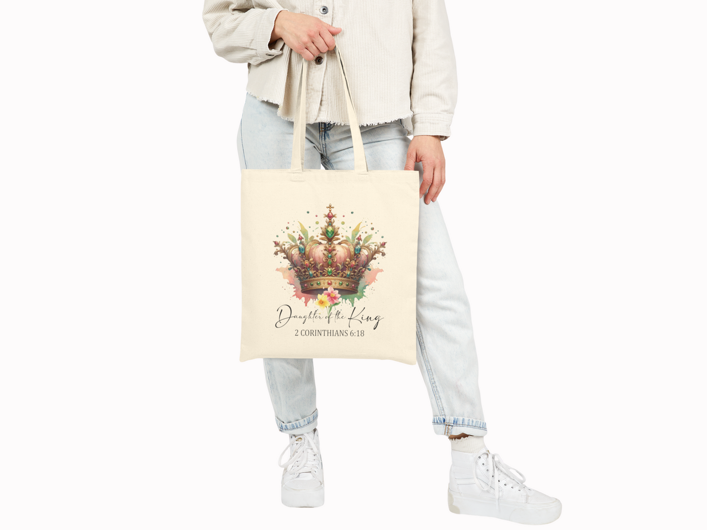 A person holds a cream tote bag featuring a colorful crown design with floral accents. Text below reads "Daughter of the King, 2 Corinthians 6:18." The tone is elegant.