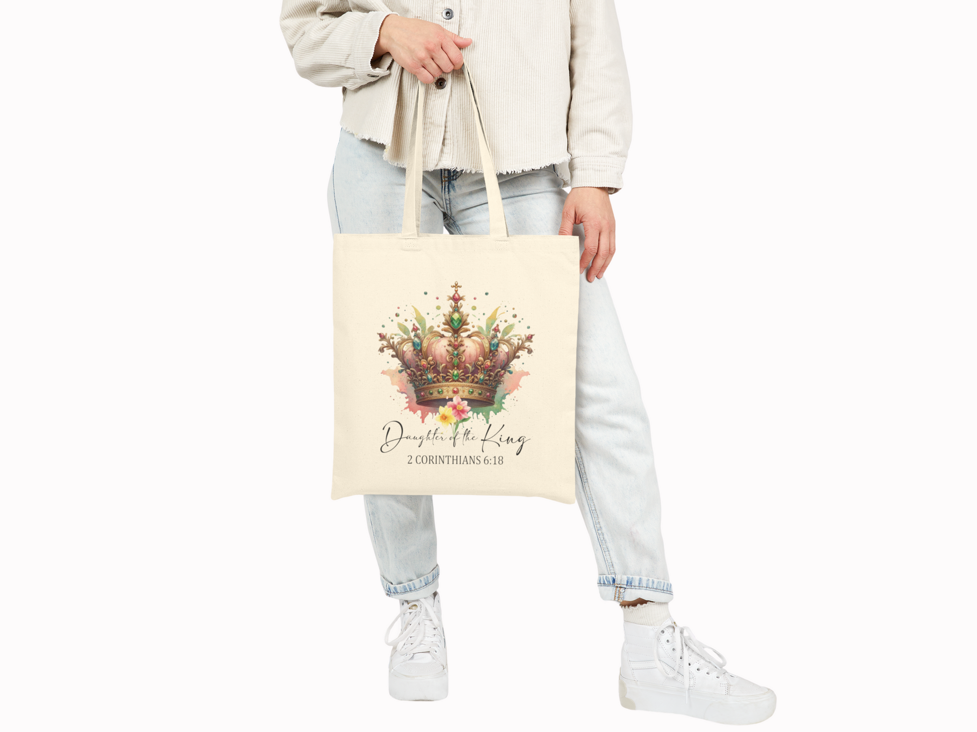 A person holds a cream tote bag featuring a colorful crown design with floral accents. Text below reads "Daughter of the King, 2 Corinthians 6:18." The tone is elegant.