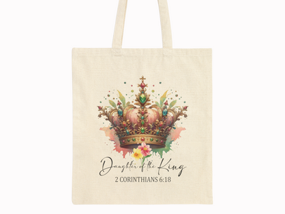 A beige tote bag features a vibrant, ornate crown with jewels and floral elements. Below, it reads "Daughter of the King, 2 Corinthians 6:18" in elegant script.