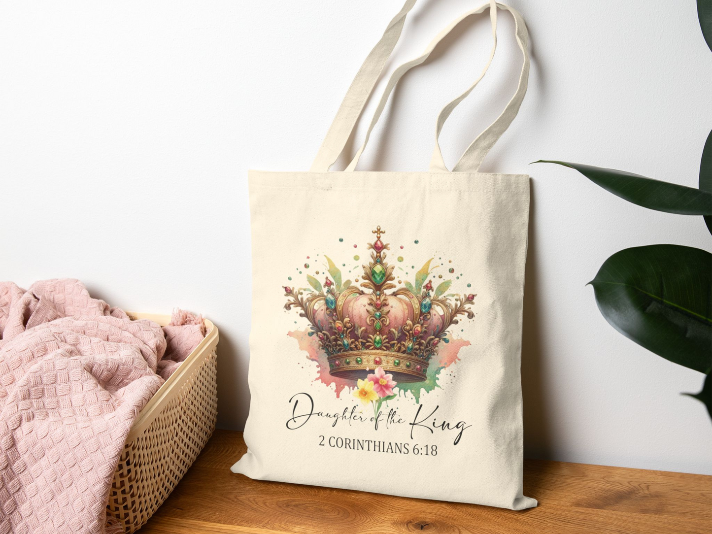 A beige tote bag with a colorful crown design and flowers, displaying "Daughter of the King, 2 Corinthians 6:18." It's on a wooden table near a pink blanket and green plant.