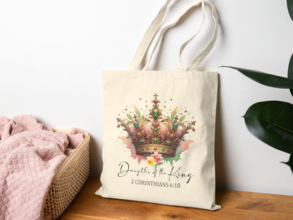 A beige tote bag with a colorful crown design and flowers, displaying "Daughter of the King, 2 Corinthians 6:18." It's on a wooden table near a pink blanket and green plant.