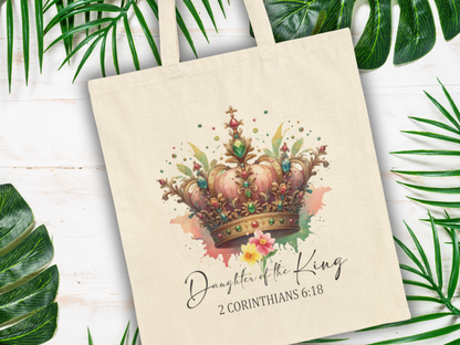 Tote bag with a colorful crown design, floral accents, and the phrase "Daughter of the King, 2 Corinthians 6:18" against a white wood background with green leaves.