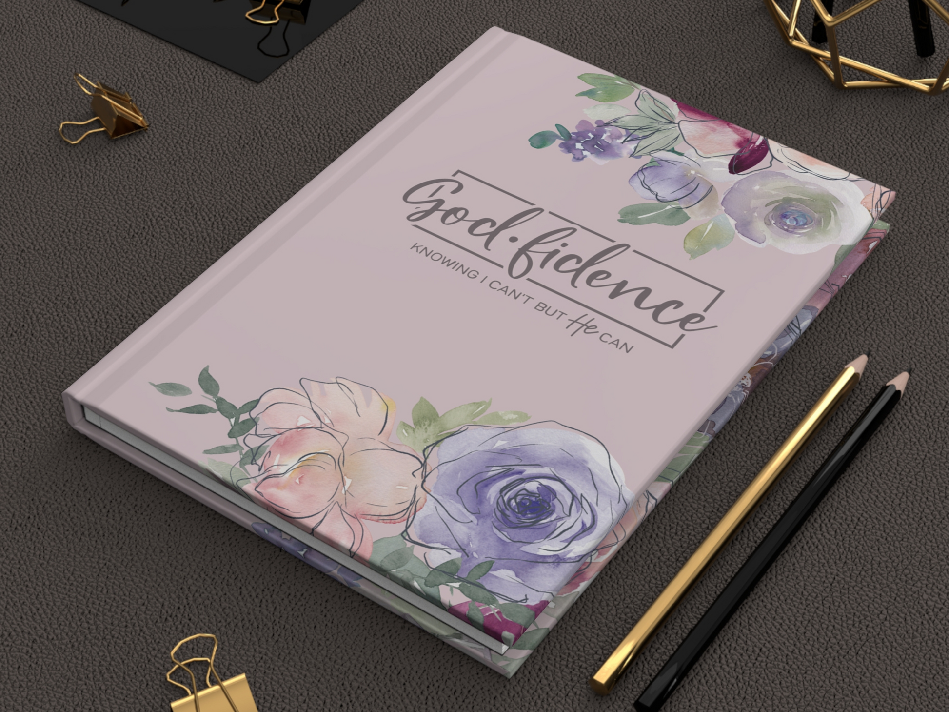 A pastel purple journal with floral designs and "Godfidence: Knowing I Can't But He Can" on the cover, placed on a textured surface beside gold and black pencils.