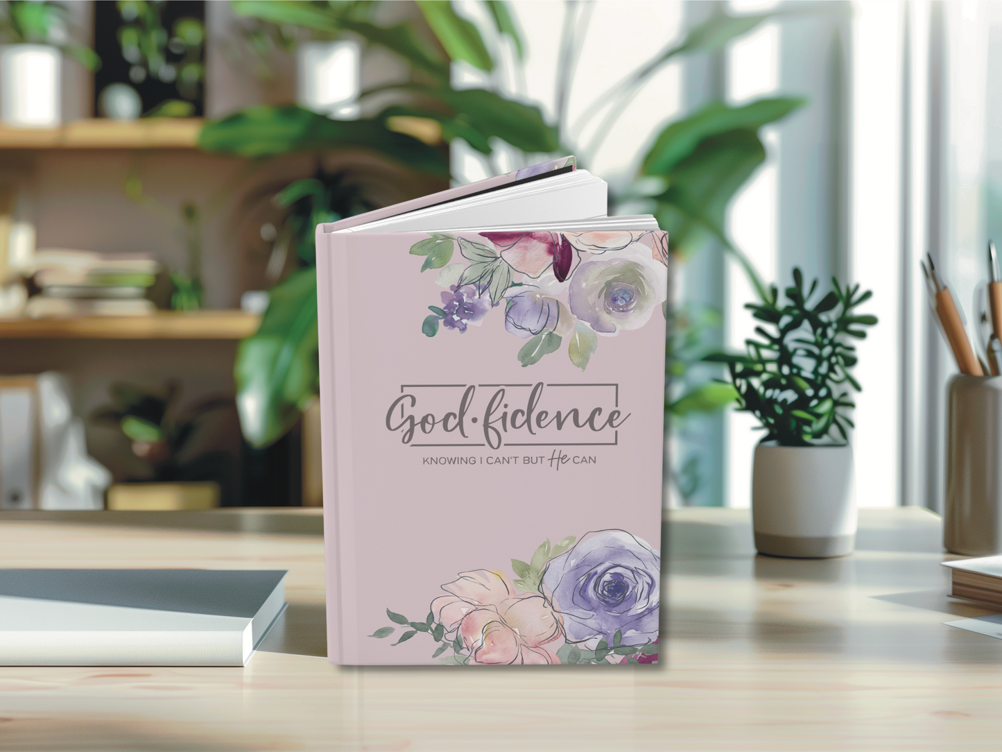 A floral-themed book titled "Godfidence" stands on a sunlit wooden desk. The cover features pastel flowers, evoking a calm and inspirational mood.