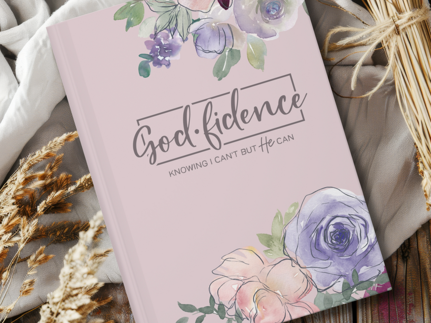 A purple notebook with watercolor flowers framing "Godfidence: Knowing I Can't But He Can" on the cover, resting on a cozy blanket beside dried wheat.
