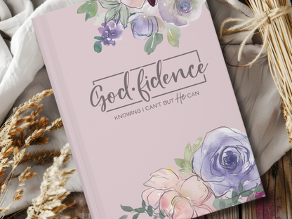 A purple notebook with watercolor flowers framing "Godfidence: Knowing I Can't But He Can" on the cover, resting on a cozy blanket beside dried wheat.
