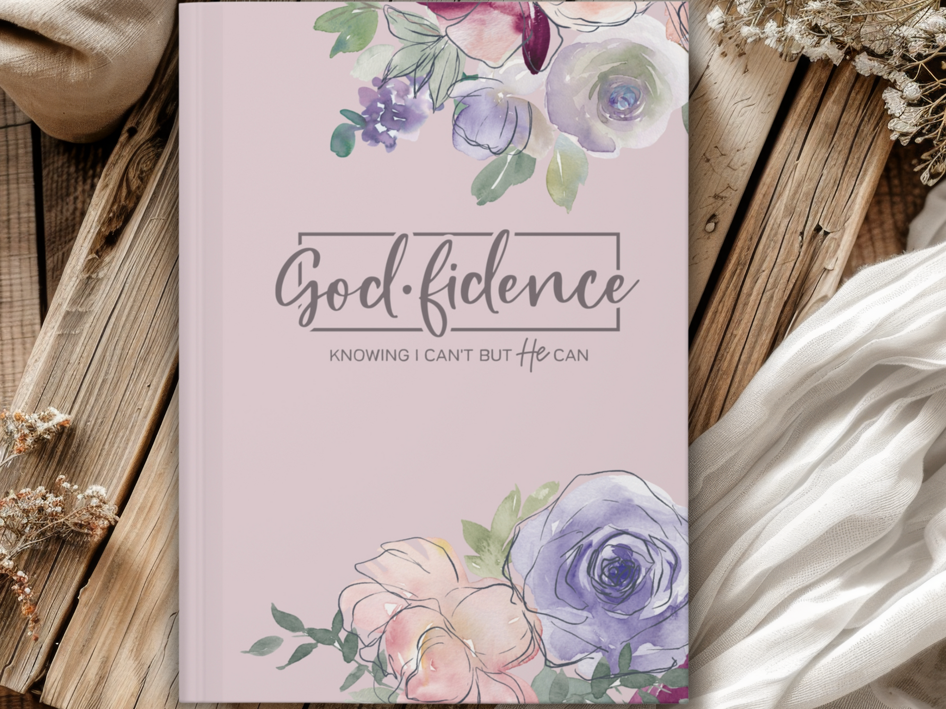 Purple journal with floral watercolor accents features text "God-fidence: Knowing I can't but He can." It rests on a wooden surface with fabric and dried flowers, evoking a serene, reflective mood.