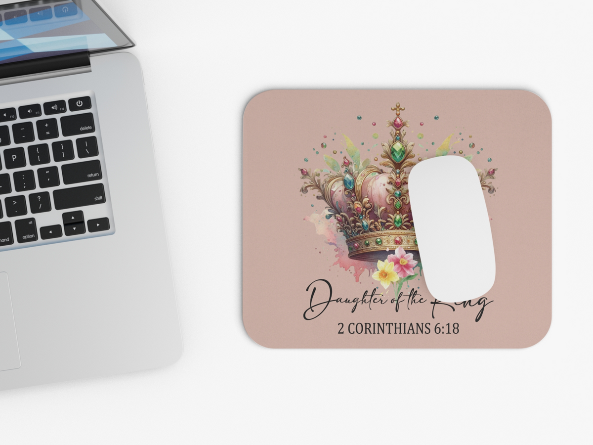 A laptop and a mouse pad on a white surface. The mouse pad features a jeweled crown with flowers and text: "Daughter of the King, 2 Corinthians 6:18."