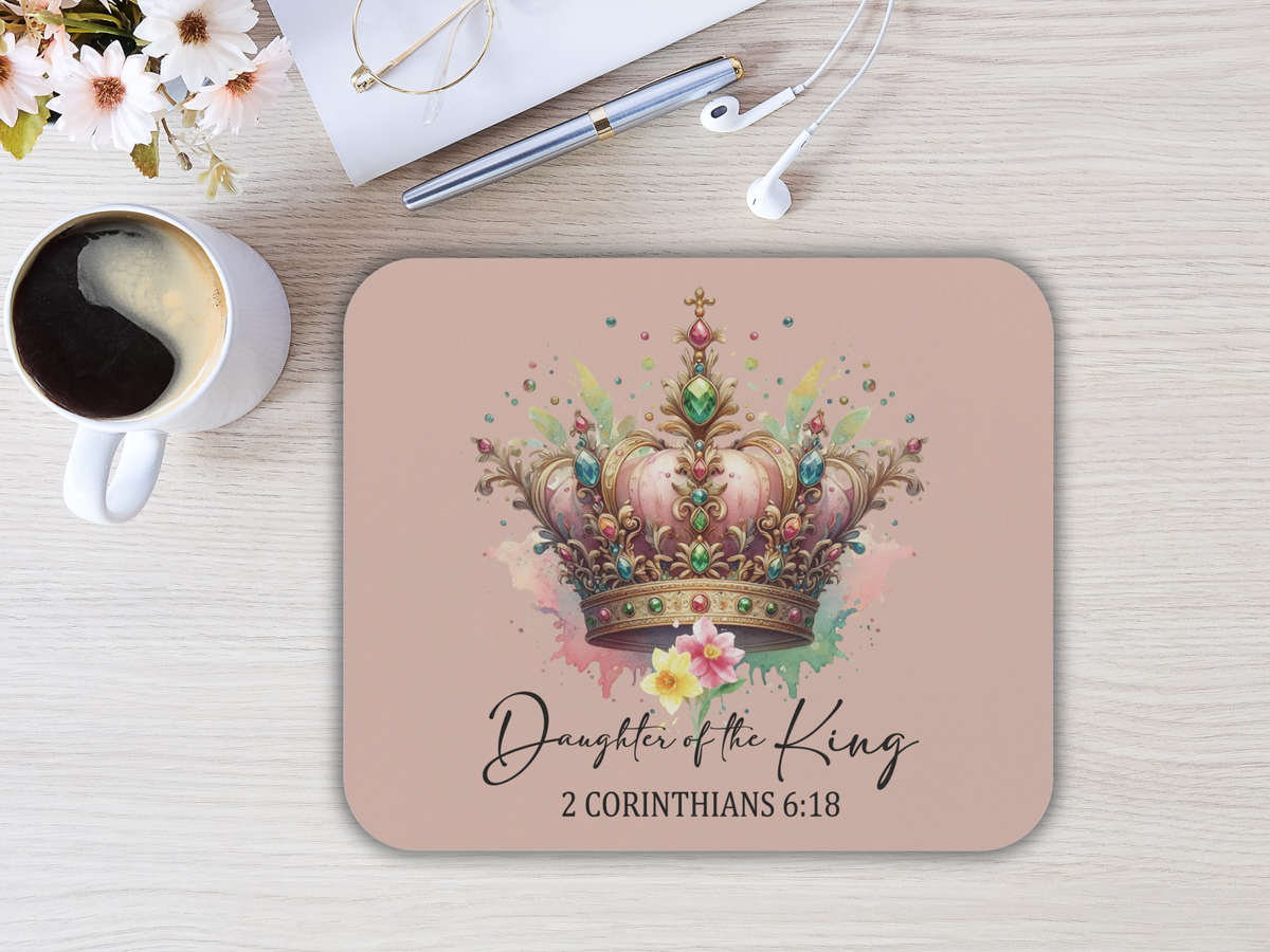 A mouse pad with floral crown and colorful jewels is on a beige background with text "Daughter of the King, 2 Corinthians 6:18." Nearby, there's a coffee cup, flowers, glasses, a pen, and earphones on a light wood desk. The scene conveys elegance and spirituality.