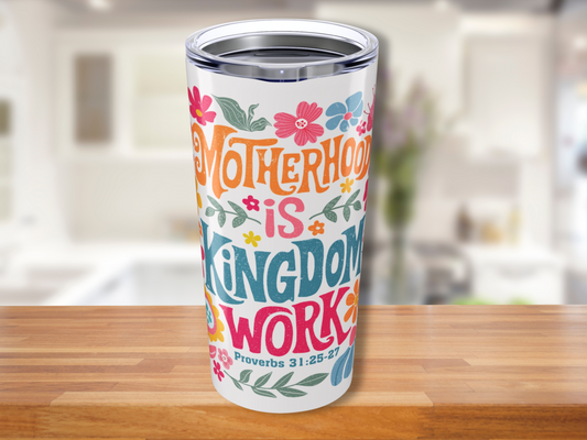 Tumbler on a wooden table with colorful text: "Motherhood is Kingdom Work. Proverbs 31:25-27" surrounded by floral designs, in a bright kitchen setting.