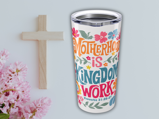 A colorful tumbler with floral designs and the text "Motherhood is Kingdom Work, Proverbs 31:25-27," beside a wooden cross and pink flowers.