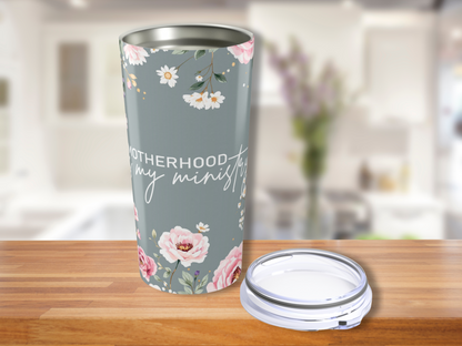A floral travel mug with the words "Motherhood is my ministry" sits on a wooden counter. The background shows a blurred kitchen setting, adding a warm, homely feel.